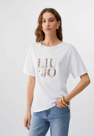 Nő fehér "LIU JO" feliratú díszített pólóban, kék farmerben, arany fülbevalóval és arany karkötőkkel, semleges háttér előtt.