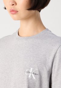 Pilka apvalaus kaklo marškinėliai, pagaminti iš medvilnės mišinio, su balta „Calvin Klein“ logotipo išsiuvinėjimu kairėje krūtinės pusėje. Lygus audinys, kasdieniškas dizainas.