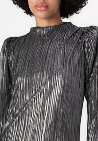 Metalni srebrni dugosleeve top s vertikalnim nabranima, visokim ovratnikom i nabranim ramenima. Glatka tekstura s reflektivnom završnicom.