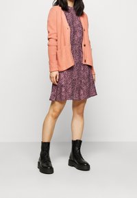 Růžový pletený kardigan přes fialové šaty s leopardím potiskem, doplněný černými šněrovacími botami. Šaty mají rozšířenou sukni a krátké rukávy.