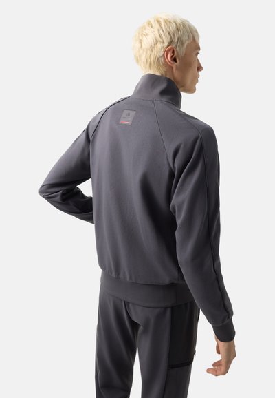 Persoon met kort blond haar, gekleed in een donkergrijs jack met rits en bijpassende broek, met de rug naar voren tegen een effen lichte achtergrond.