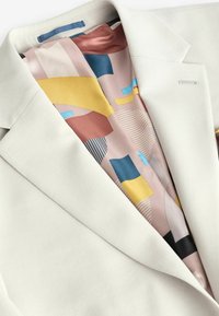 Blazer de color claro con solapas puntiagudas, que presenta un forro de satén estampado en diseños geométricos en tonos rosa, azul, amarillo y negro.
