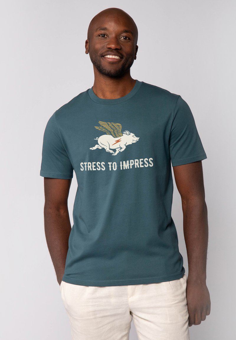 T-shirt en coton teal avec un graphique blanc d'un cochon volant et la phrase "STRESS TO IMPRESS" en lettres grasses. Manches courtes, coupe décontractée.