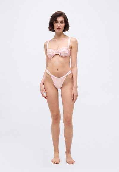 Γυναίκα που στέκεται ξυπόλητη μπροστά σε λευκό φόντο, φορώντας ανοιχτό ροζ μαγιό με φαρδιές τιράντες και σλιπ με ψηλή κοπή.