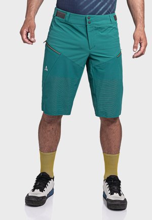 Mann in türkisfarbenen knielangen Shorts mit Reißverschlusstaschen, gelben Socken und Schwarz-Weiß-Sneakern, stehend vor einem weißen Hintergrund.