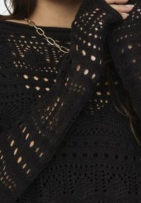 Pull noir en maille ajourée avec motifs géométriques porté avec un collier chaîne dorée, montrant un gros plan du haut du torse et du bras.