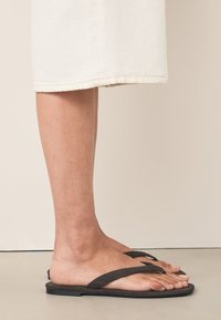 Sorte flade sandaler med to krydsede remme, lavet af blødt materiale, der viser et minimalistisk design. Båret på en bar fod, uden bemærkelsesværdige mønstre.