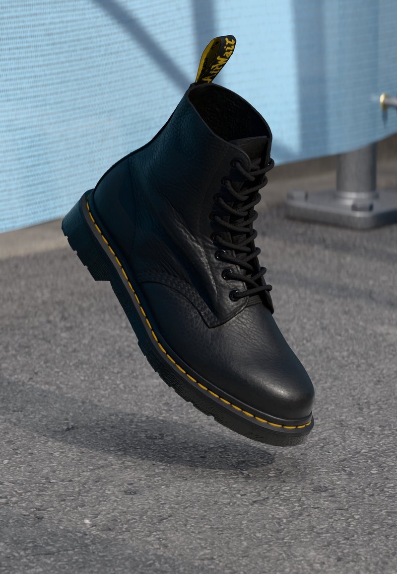Botín negro de cuero hasta el tobillo con costuras amarillas, diseño de cordones y un tirador en el talón. Presenta una parte superior texturizada y una suela resistente.