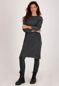 Vrouw met lang bruin haar die een donkergrijze lange mouwen gebreide jurk met studs, zwarte leren leggings en zwarte veterschoenen draagt, poseert met gekruiste armen.