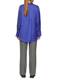 Satijnen blauwe blouse met een kraag en lange mouwen, met zijsplitten. Gepaard met lichtgrijze, op maat gemaakte pantalons en zwarte schoenen.