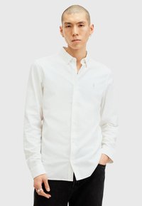 Hvid langærmet skjorte med button-down krave, diskret logo-detail og glat tekstur, parret med mørk denim jeans.