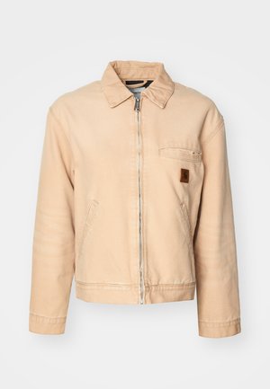 Beige denimjacka med dragkedja fram, spetsig krage, två sidofickor och en liten logopatch på bröstet. Avslappnad, lös passform.