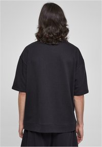 Černá oversize mikina vyrobená z měkkého materiálu, s kleslými rameny, krátkými rukávy a rovnými lemy. Žádné viditelné logo ani vzory.