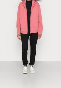 Osoba v růžové zipové mikině, černém topu a kalhotách, a bílých teniskách stojící na světlém jednobarevném pozadí.