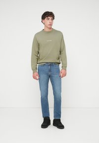 Πράσινο φούτερ με ριμπ λαιμόκοψη, με λευκό κείμενο. Μπλε τζιν με ίσια γραμμή και μαύρα αθλητικά παπούτσια. Απλός σχεδιασμός και καθημερινή ένδυση.