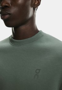 Zielony bawełniany sweter z okrągłym dekoltem, z czarnym logo na piersi. Tkanina wydaje się gładka o miękkiej fakturze.