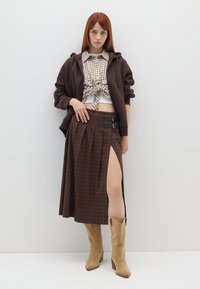 Sudadera marrón, camisa de cuadros anudada a la cintura, falda maxi de cuadros marrón con abertura en el muslo y botas de cowboy de ante color arena.
