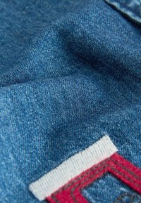 Tessuto denim blu con una trama strutturata. Presenta un'etichetta bianca e rossa con dettagli di cucitura, parzialmente visibile nell'angolo inferiore.