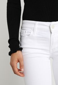 Jeans ajustados blancos hechos de denim, con cierre frontal de botón y bolsillos laterales. Llevados con una blusa negra de manga larga de canalé.