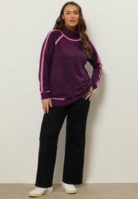 Lila Rollkragenpullover mit rosa Seitenstreifen, locker sitzend. Kombiniert mit schwarzen weiten Hosen und weißen Sneakern.
