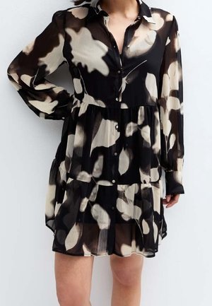 Femme portant une robe noire à manches longues boutonnée avec de grands motifs floraux abstraits beiges et blancs, debout avec une main sur la hanche.