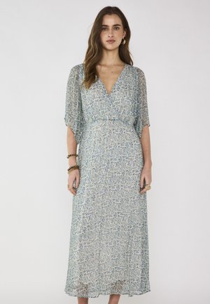 Donna con capelli lunghi e castani, indossa un abito midi azzurro chiaro a fiori con maniche corte a volant e orecchini a cerchio dorati, in piedi davanti a uno sfondo neutro.