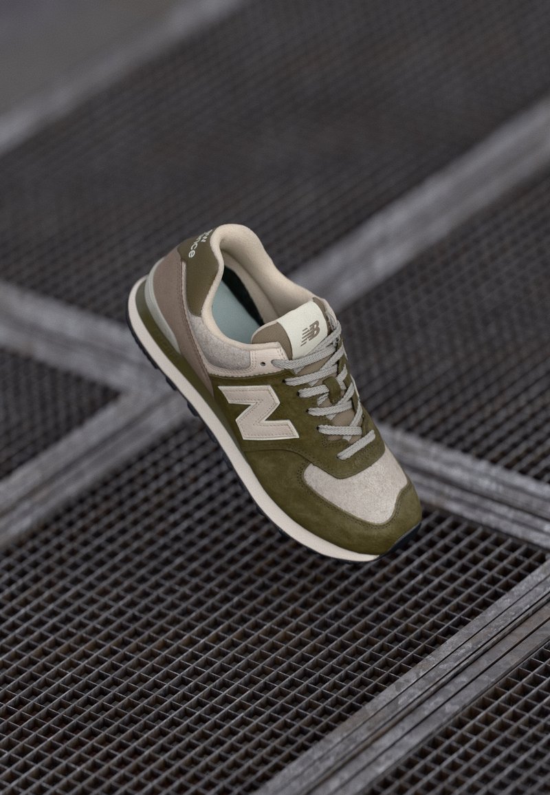 Baskets New Balance vert olive et beige avec logo blanc « N », lacets gris, reposant sur un sol en grille métallique.