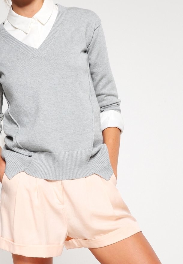 Persoon in een wit overhemd met kraag, een lichtgrijze V-halstrui en een lichtroze korte broek met de handen in de zakken tegen een effen achtergrond.