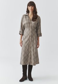 Vestito lungo a quadri nei toni beige e marrone, con chiusura frontale a bottoni, colletto e maniche arrotolate, abbinato a stivali al ginocchio in suede marrone.