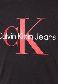 Svart T-shirt med stor röd och vit text: "Calvin Klein Jeans" med en framträdande "CK" logotyp i röd färg på framsidan.