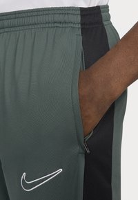Nike sportbroek heeft een groene stof met zwarte zijkanten, een elastische tailleband, ritszakken en witte logo-details.