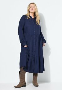 Marineblauwe knoopjurk met lange mouwen, gelaagd ontwerp en zakken; gecombineerd met bruine cowboylaarzen met decoratieve stitchings.