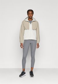 Ελαφρύ αδιάβροχο δίχρωμο μπουφάν, με μπεζ επάνω μέρος και λευκή κάτω επιφάνεια, με κουκούλα και ελαστικά μανίκια. Συνδυάζεται με γκρι κολάν και αθλητικά παπούτσια.