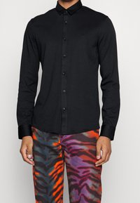 Camicia nera a maniche lunghe con bottoni e collo classico, caratterizzata da un design elegante e un branding discreto. Indossata con pantaloni a fantasia multicolore.