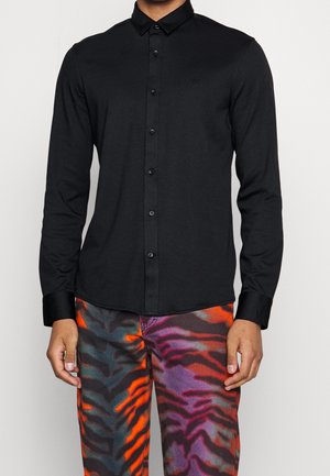 Black long-sleeve button-up shirt with a classic collar, featuring a sleek design and subtle branding. Worn with multicolored patterned pants.