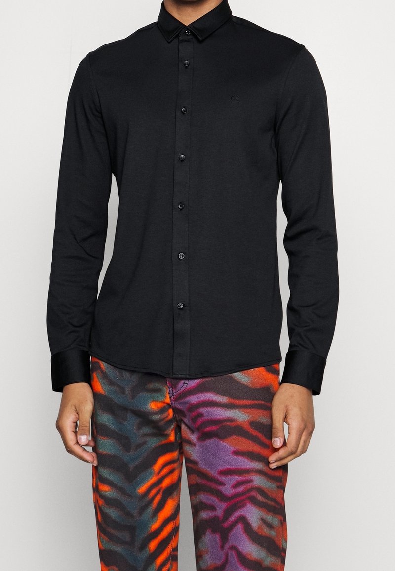 Camicia nera a maniche lunghe con bottoni e collo classico, caratterizzata da un design elegante e un branding discreto. Indossata con pantaloni a fantasia multicolore.