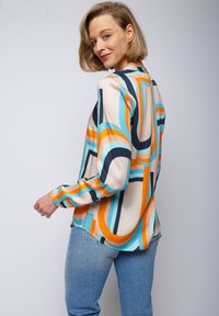 Langsleeve blouse met een gekleurd geometrisch patroon in oranje, blauw en marine op een beige achtergrond, gemaakt van een gladde stof.