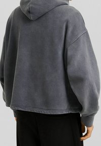 Felpa grigia corta con tessuto strutturato, dotata di cappuccio e maniche larghe e comode. L'orlo si trova appena sopra la linea della vita.