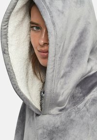 Couverture à capuche grise en tissu doux et pelucheux avec une doublure blanche duveteuse. Elle présente une grande capuche et une coupe décontractée.