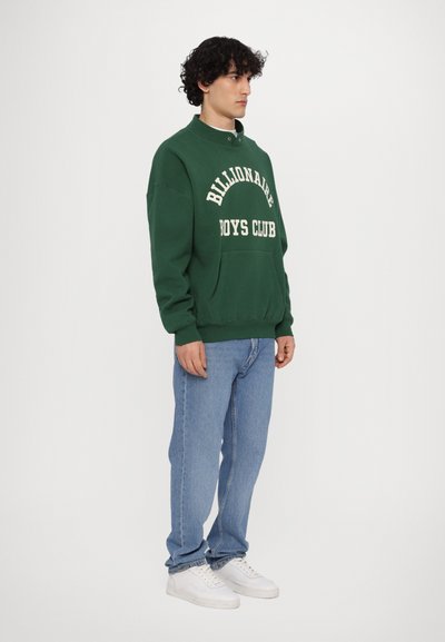 Sweatshirt vert avec le texte « Billionaire Boys Club » en blanc, associé à un jean bleu clair et des baskets blanches. Coupe standard et col rond.