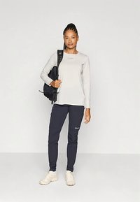 Lichtgrijs shirt met lange mouwen en logo, gecombineerd met donkerblauwe pantalon. Model draagt een zwarte rugzak en crèmekleurige sneakers.