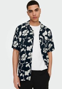 Kurzärmliges, marineblaues Hemd mit großen weißen Blumenmustern, offenem Kragen und Knöpfen. Über einem weißen T-Shirt mit lockerer Passform getragen.
