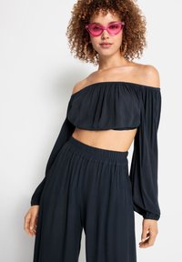 Marinblå av-axeln crop top med långa ärmar, kombinerad med vida byxor. Accessoarer inkluderar rosa kattögon-solglasögon och stora hoopsörhängen.