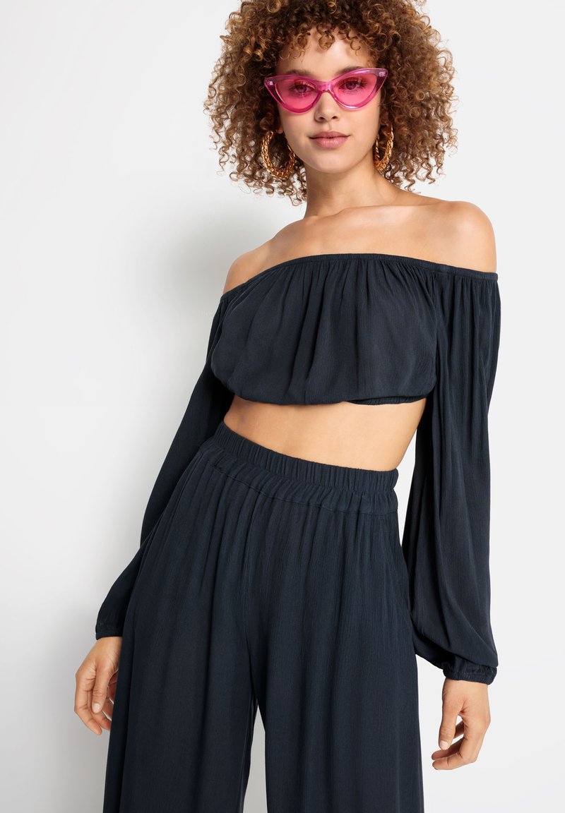 Marinblå av-axeln crop top med långa ärmar, kombinerad med vida byxor. Accessoarer inkluderar rosa kattögon-solglasögon och stora hoopsörhängen.