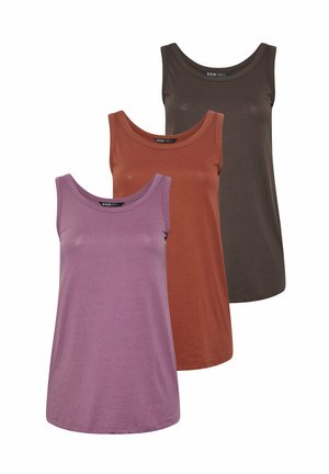 Kvinde med blondt hår iført en ærmeløs lilla top, lyseblå skinny jeans og hvide sneakers, stående mod en ensfarvet baggrund.