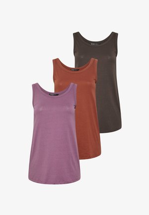 Kobieta o blond włosach, ubrana w fioletowy top bez rękawów, jasnoniebieskie obcisłe jeansy i białe trampki, stojąca na jednolitym tle.