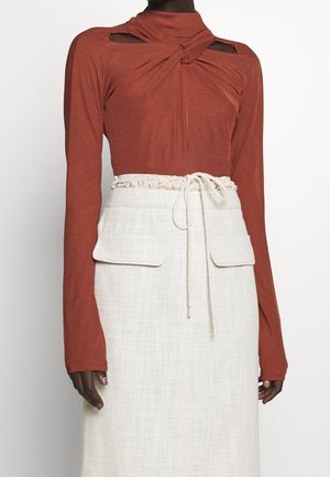 Haut à manches longues couleur rouille avec encolure torsadée et découpes, associé à une jupe crème taille haute dotée d'un cordon de serrage et de poches à rabat.