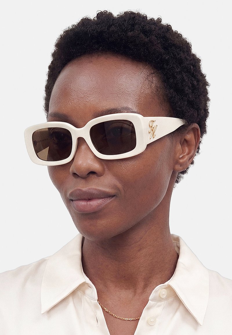 Personne avec des cheveux courts et bouclés portant de grandes lunettes de soleil blanches rectangulaires et une chemise blanche à col avec un fin collier en or.