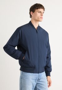 Veste de bombardier marine avec col montant, fermeture éclair sur le devant, poches latérales et poignets côtelés. Tissu lisse et léger. Design classique.