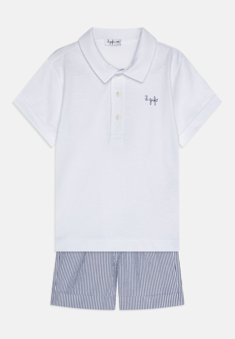 Weißes Poloshirt mit Kragen und drei Knöpfen, versehen mit einem maritimen Logo. Kombiniert mit kurzärmeligen blau-weißen gestreiften Shorts.